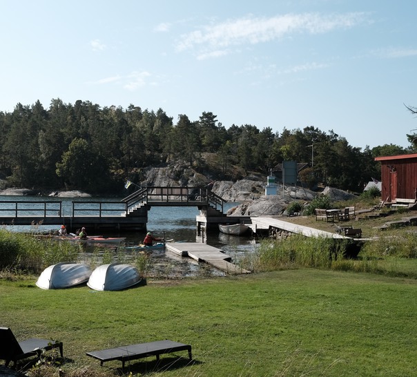 Skärgårdslandskap med vatten, bryggor, båtar och solstolar.