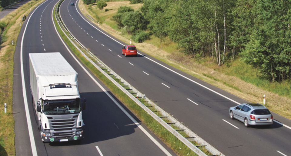 Motorväg med vit lastbil körandes på vägen och två personbilar körandes i motsatt riktning. Runt vägen syns grön sko