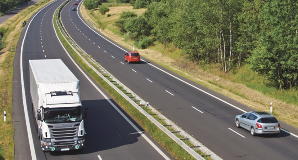 Motorväg med vit lastbil körandes på vägen och två personbilar körandes i motsatt riktning. Runt vägen syns grön sko