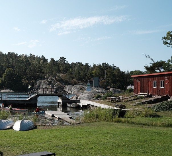 Vy över gräsplan, vatten, båtar och röd stuga.