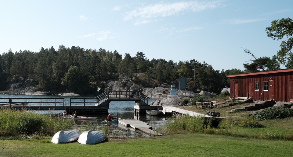 Vy över gräsplan, vatten, båtar och röd stuga.