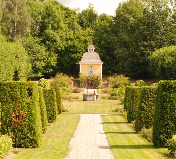En grusgång i en symmetrisk trädgård kantad av formklippta buskar som leder fram till en brunn.