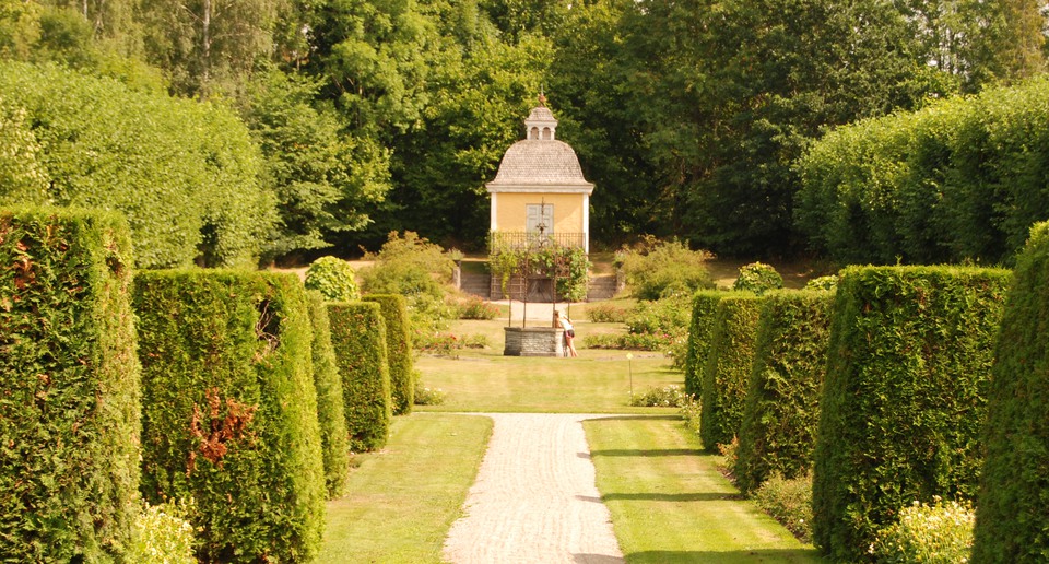 En grusgång i en symmetrisk trädgård kantad av formklippta buskar som leder fram till en brunn.