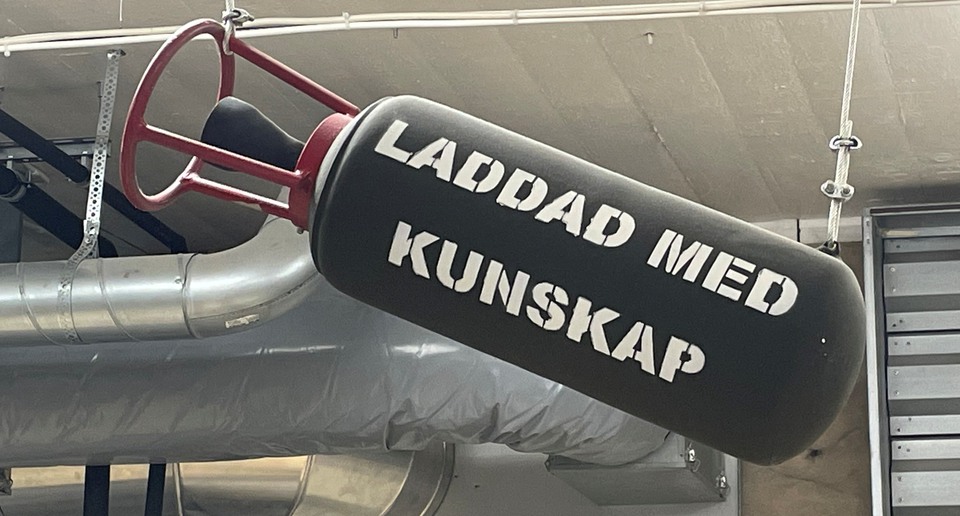 Svart hängande boxningssäck med texten "Laddad med kunskap" i en industriliknande miljö.