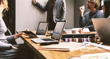 Personer sitter vid ett bord med datorer, en står vid en whiteboard-tavla.