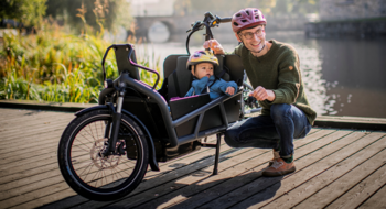 En person med cykelhjälm sitter på huk bredvid en lådcykel med ett barn som också har hjälm.