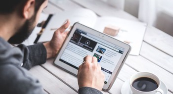 En person sitter vid ett bord och läser på en surfplatta med en kopp kaffe.