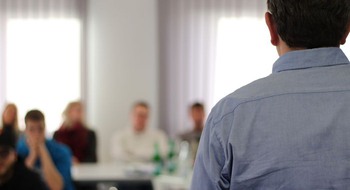 Person pratar framför ungdomar som sitter vid bord. 