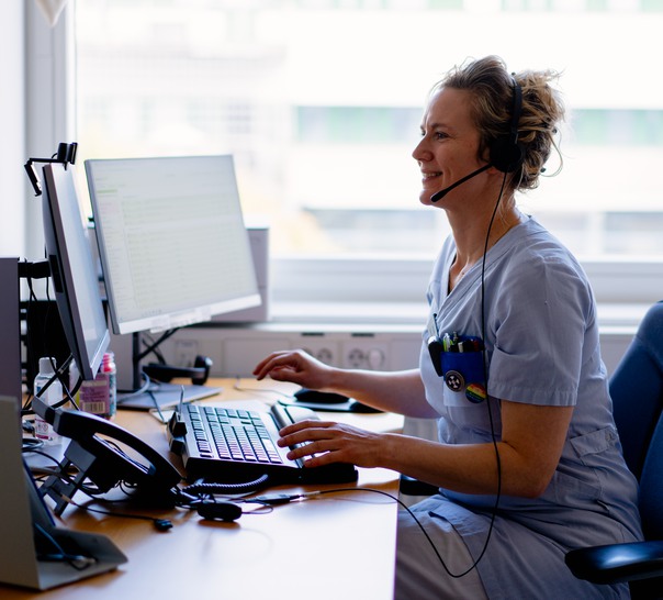 En person i vårdkläder sitter vid en dator med headset.